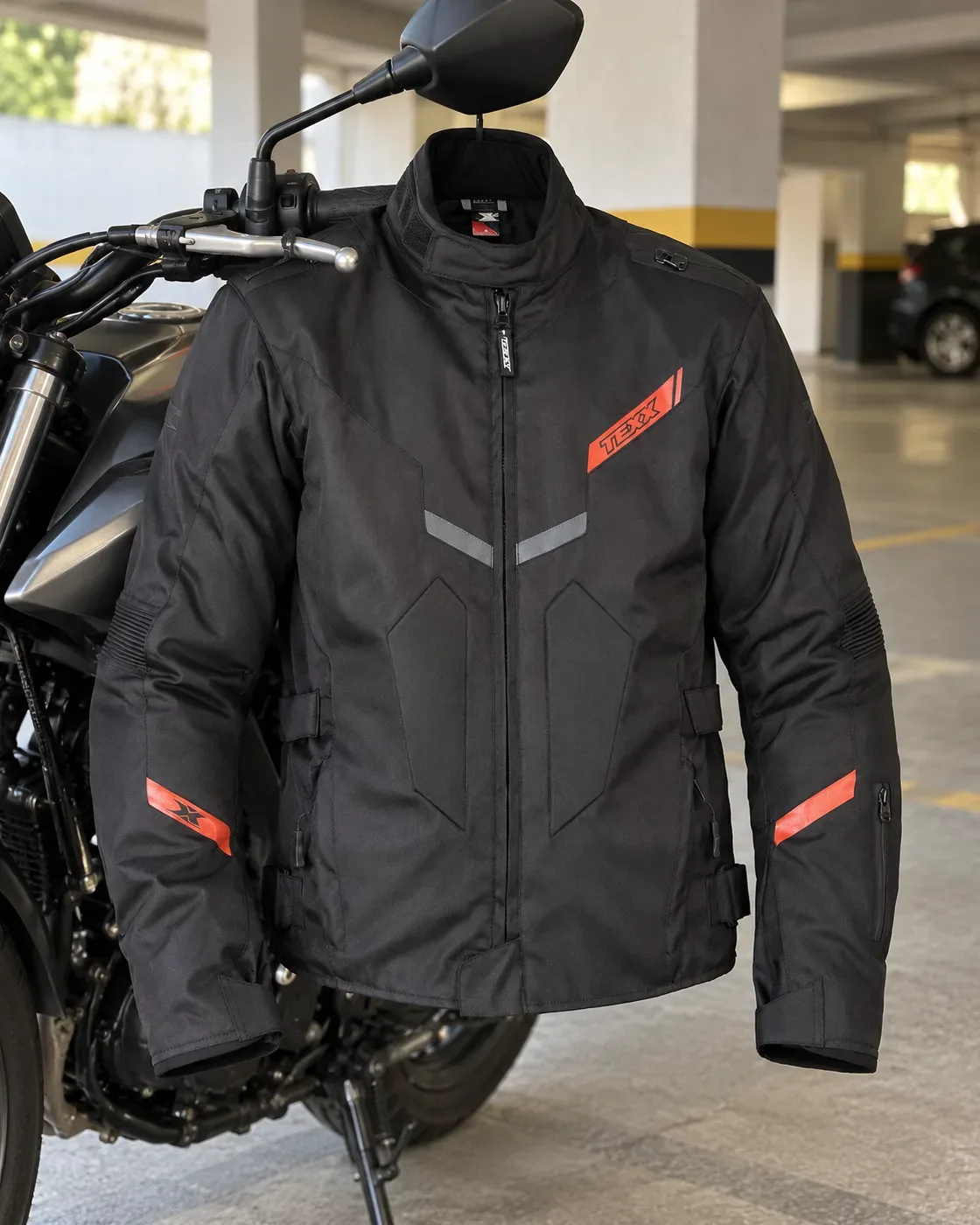 Jaqueta Texx New Strike V3 preta, com faixas vermelhas no peito e nos braços, pendurada no retrovisor de uma moto numa garagem de prédio